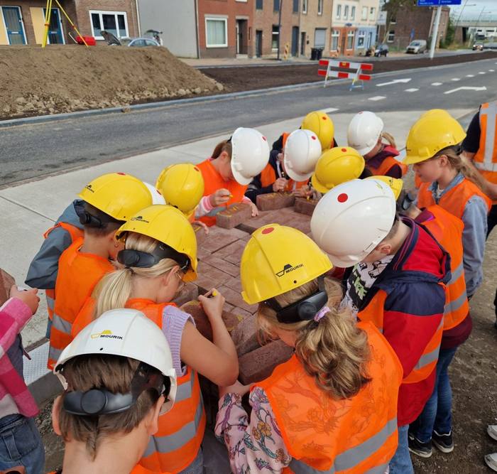 Schoolkinderen met helm en hesje schrijven hun naam op stenen bij de bouwplaats